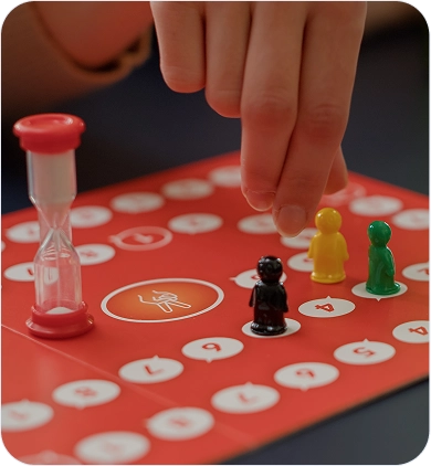 Hand die een speelfiguur verplaatst op een rood bordspel met zandloper en gekleurde pionnen.